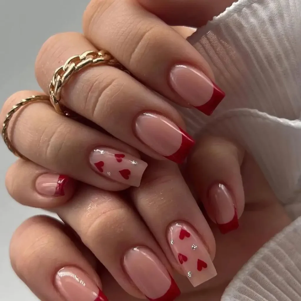 Close-up of manicured nails featuring pink base with red French tips and accent nails decorated with small red hearts and silver glitter details. Two gold chain rings visible on fingers against light gray background.