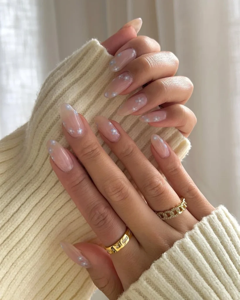 Almond-shaped clear glossy nails photographed against a cream knit sweater, with delicate gold rings adorning the fingers.