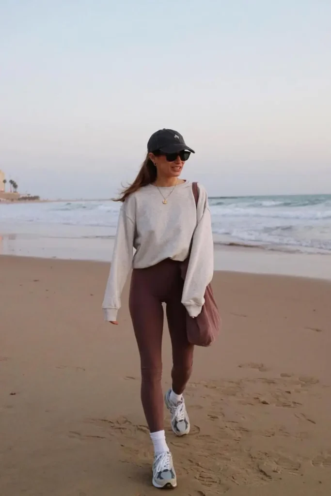 Cozy neutral-toned outfit with a cream sweatshirt, chocolate brown leggings, and white sneakers. Ideal for breezy beach days, offering a stylish yet laid-back aesthetic.