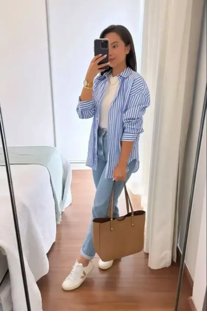 A blue and white striped button-down shirt layered over a white tee with light wash jeans for a timeless spring look. A tan tote bag adds warmth, while white sneakers ensure all-day comfort. Perfect for casual Fridays or weekend errands.