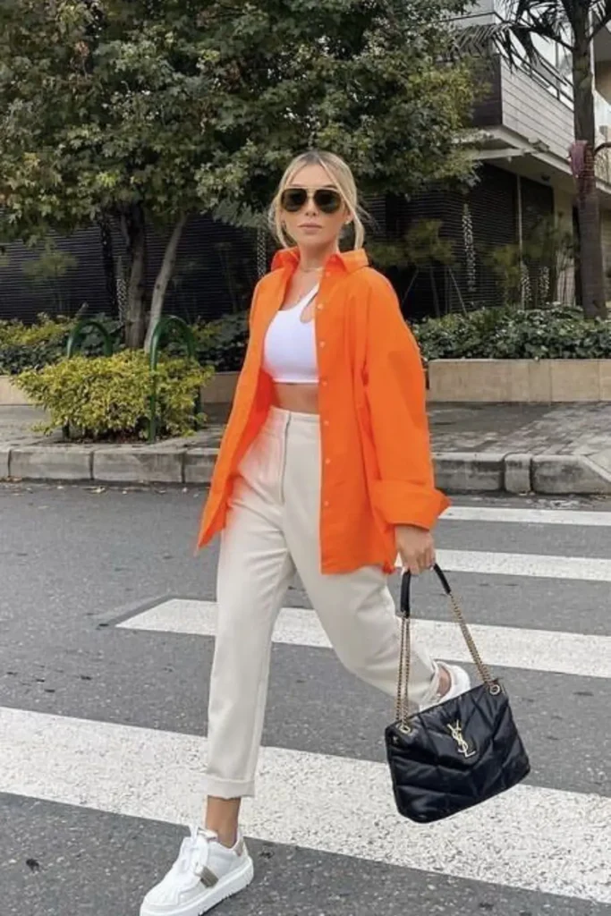 A striking orange oversized blazer paired with a white crop top, high-waisted cream trousers, and white sneakers. The structured black handbag adds sophistication, making this outfit a bold yet wearable choice for stylish city looks.