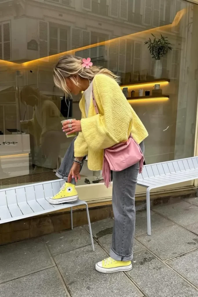 Vibrant street style featuring a bright yellow oversized sweater, gray jeans, and matching yellow sneakers. The pink shoulder bag and floral hair accessory add playful charm. Photographed outside a cozy coffee shop.