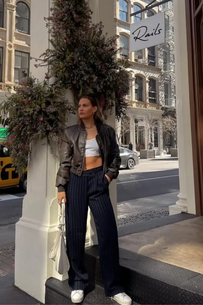 Trendy urban fashion with a black leather jacket, white crop top, and navy pinstriped pants. Paired with white sneakers and a white bag, this effortlessly cool look.