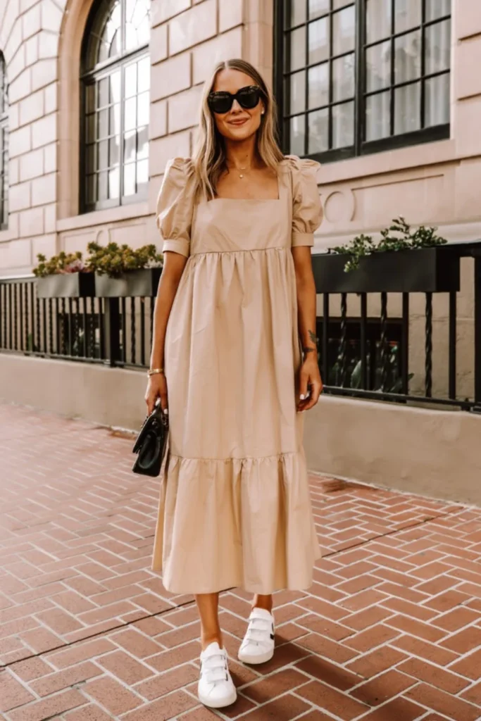 A neutral beige midi dress with puff sleeves and a tiered skirt, featuring a square neckline and empire waist for a flattering silhouette. Paired with white sneakers and a small black handbag, this outfit combines casual comfort with sophistication, making it perfect for summer date nights, gallery visits, or upscale shopping trips.