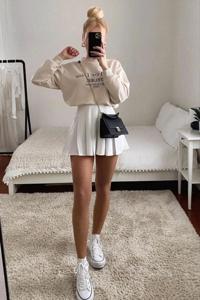 This outfit embraces the tennis-inspired trend with a cream sweatshirt and a pleated white mini skirt, creating a clean and preppy aesthetic. The white high-top sneakers enhance the sporty vibe, while a small black crossbody bag adds a touch of elegance. Perfect for transitioning from casual daytime activities to evening hangouts, this look is both playful and sophisticated.