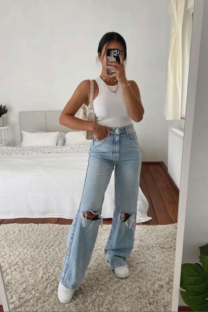 A classic white tank top styled with light wash wide-leg jeans featuring trendy knee rips. White sneakers and a small shoulder bag complete this effortlessly fashionable mirror selfie outfit.