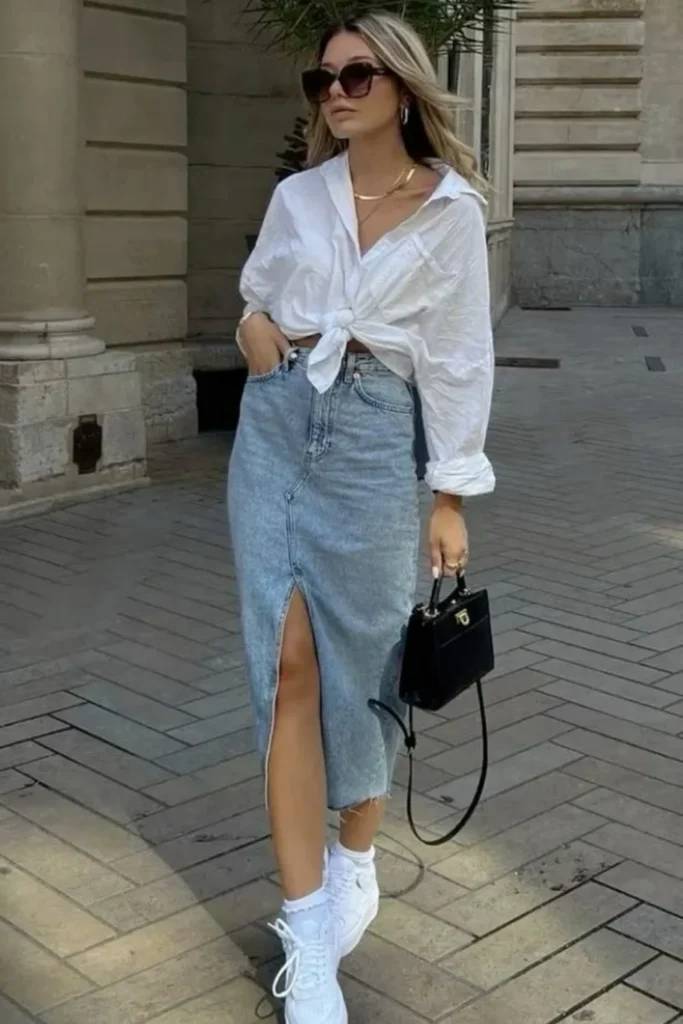 A light-wash denim midi skirt with a front slit, paired with an oversized white button-up tied at the waist. Accessorized with sunglasses, a gold necklace, and a black mini handbag, this outfit is completed with crisp white sneakers. Perfect for brunch or gallery hopping, offering a put-together yet comfortable look.